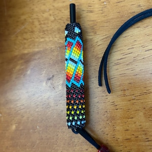 Peut inclure: Un bouchon de bouteille perlé avec une tige noire et un motif perlé coloré. La section perlée présente un motif de perles rouges, oranges, jaunes, turquoise et noires. Un cordon noir est attaché au bouchon.