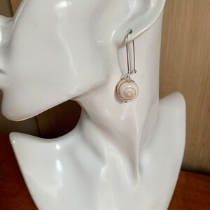 May include: A pair of silver dangle earrings with a white seashell pendant. The seashell has a brown and white swirl pattern.