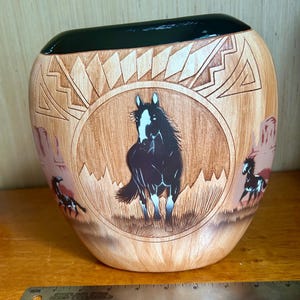 May include: A ceramic vase with a black top and a light brown body. The vase features a central image of a black horse with a white blaze, surrounded by a circular design. Additional horse figures are depicted on the sides.