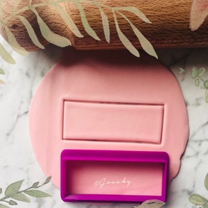 May include: A purple plastic rectangle cookie cutter with the text "@gouaky" inside. The cutter is on a pink surface with a rectangular impression in the surface.