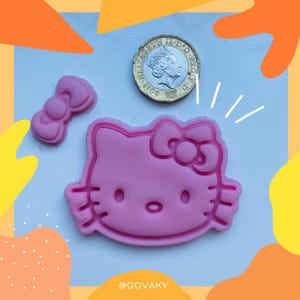 May include: Pink plastic cookie cutter shaped like Hello Kitty with a bow on top. The cookie cutter is on a white surface.