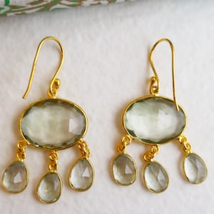 May include: A pair of gold earrings with a teardrop-shaped green gemstone pendant. The earrings have a dangling design with three smaller teardrop-shaped gemstones.