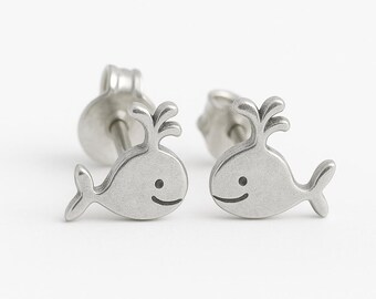 May include: A pair of silver-colored whale stud earrings. Each earring features a stylized whale design with a smiling face and a water spout. The earrings are set against a white background.