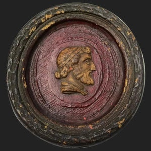 May include: A round wooden plaque with a raised image of a man's head in profile. The plaque is painted red and has a dark brown border.