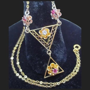 May include: A gold necklace with a geometric design featuring two triangular pendants with floral accents. The pendants are adorned with red and white gemstones.