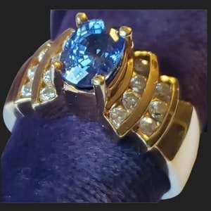 May include: A gold ring with a large blue sapphire gemstone surrounded by smaller diamonds. The ring has a unique design with the diamonds set in a curved pattern.
