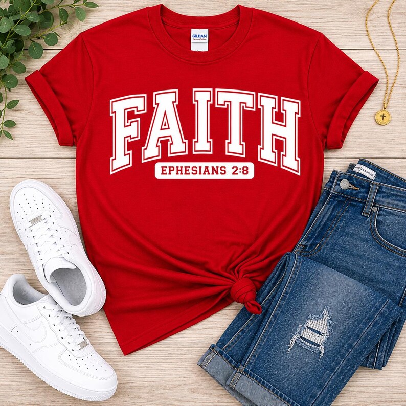 Puede incluir: Una camiseta roja de manga corta con la palabra FAITH en letras blancas grandes y el texto EPHESIANS 2:8 impreso debajo. La camiseta se combina con pantalones vaqueros azules y zapatillas blancas para un conjunto informal.