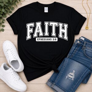 Puede incluir: Una camiseta negra de manga corta que presenta la palabra FAITH en letras blancas grandes de estilo universitario, con el texto EPHESIANS 2:8 impreso en un recuadro blanco m&aacute;s peque&ntilde;o justo debajo de la palabra principal en la parte frontal.