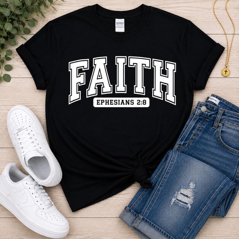 Puede incluir: Una camiseta negra de manga corta que presenta la palabra FAITH en letras blancas grandes de estilo universitario, con el texto EPHESIANS 2:8 impreso en un recuadro blanco m&aacute;s peque&ntilde;o justo debajo de la palabra principal en la parte frontal.