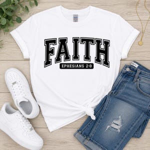 Puede incluir: Una camiseta blanca de manga corta con la palabra FAITH en letras negras grandes de estilo universitario, con el texto EPHESIANS 2:8 impreso en un recuadro rectangular negro m&aacute;s peque&ntilde;o justo debajo de la palabra principal en el frente.