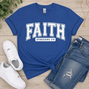 Puede incluir: Una camiseta de manga corta color azul real con la palabra FAITH en letras blancas grandes de estilo universitario, con el texto EPHESIANS 2:8 impreso en un recuadro blanco justo debajo de la palabra principal en la parte frontal.