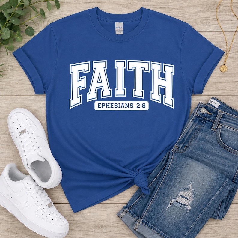 Puede incluir: Una camiseta de manga corta color azul real con la palabra FAITH en letras blancas grandes de estilo universitario, con el texto EPHESIANS 2:8 impreso en un recuadro blanco justo debajo de la palabra principal en la parte frontal.