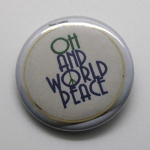 May include: A circular button featuring the text "Oh and World Peace" in green and blue. The peace symbol replaces the 'O' in 'World'. The button has a white background, a gold border, and a silver metal edge.