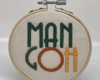 May include: Embroidered hoop art with the words "Man Goth" in green, orange, and yellow thread. The hoop is made of wood and has a metal hanging attachment.