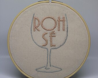 May include: Embroidered hoop art featuring the word "ROSÉ" in a stylized wine glass design. The letters are in a light pink thread, and the glass outline is in a light blue thread. The fabric is a neutral beige color, set in a wooden hoop.