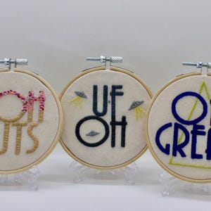 May include: Three embroidered hoops with humorous phrases. One says "DOH NUTS" with pink and brown thread. Another says "UF OH" with UFOs. The last says "OH GREAT" with a green triangle.