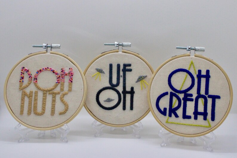 May include: Three embroidered hoops with humorous phrases. One says "DOH NUTS" with pink and brown thread. Another says "UF OH" with UFOs. The last says "OH GREAT" with a green triangle.