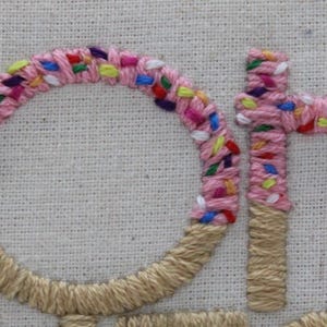 May include: Close-up of embroidered text on a cream-colored fabric. The letters "OH" are prominently displayed, with the "O" in a circle shape. The letters are pink with colorful sprinkles, and the base of the letters is a tan color.