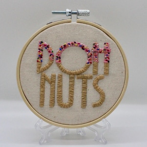 May include: Embroidered hoop art with the words "DOH NUTS" in a playful font. The letters are stitched in a light brown thread, with the "O" and the tops of the "H" and "T" decorated with colorful sprinkles. The hoop is displayed on a clear stand.