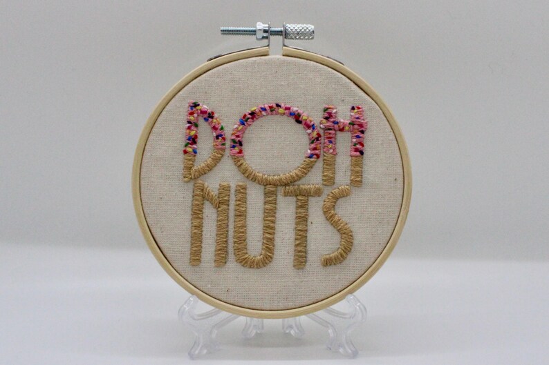 May include: Embroidered hoop art with the words "DOH NUTS" in a playful font. The letters are stitched in a light brown thread, with the "O" and the tops of the "H" and "T" decorated with colorful sprinkles. The hoop is displayed on a clear stand.