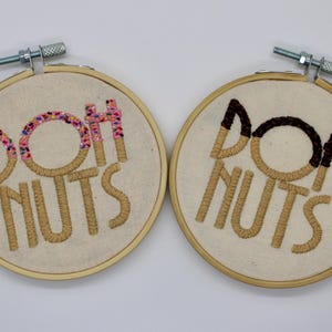 May include: Two embroidered hoops with the words "DOH NUTS". One has a pink and colorful sprinkle design on the "DOH" and "H", while the other has a chocolate-colored design on the "DOH" and "H". The embroidery is done in a tan thread on a cream-colored fabric.