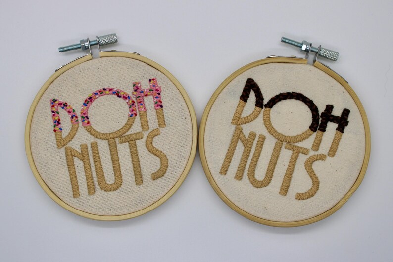 May include: Two embroidered hoops with the words "DOH NUTS". One has a pink and colorful sprinkle design on the "DOH" and "H", while the other has a chocolate-colored design on the "DOH" and "H". The embroidery is done in a tan thread on a cream-colored fabric.