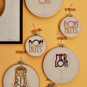 May include: A collection of embroidered hoop art pieces with food and drink-themed designs. The pieces include the words "Prosecco Oh", "Doh Nuts", "Merlot", "Burrito Oh", and "Man Coh" in various colors and fonts.