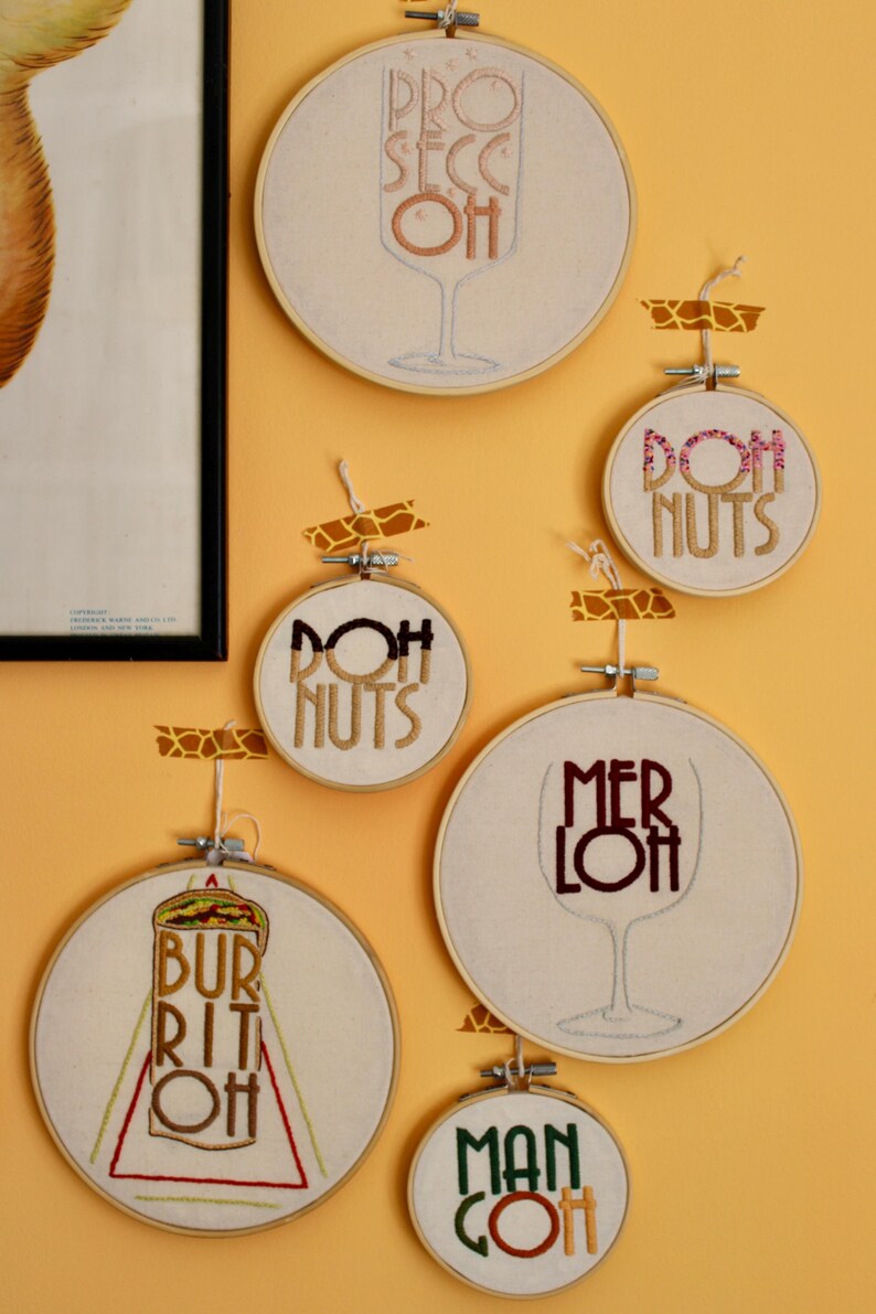 May include: A collection of embroidered hoop art pieces with food and drink-themed designs. The pieces include the words "Prosecco Oh", "Doh Nuts", "Merlot", "Burrito Oh", and "Man Coh" in various colors and fonts.