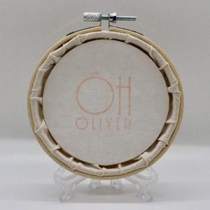 May include: Round embroidery hoop with a cream-colored fabric featuring the text "OH OLIVER" in a soft orange hue. The fabric is stretched within a wooden hoop, secured with a metal screw. The hoop is displayed on a clear plastic stand.