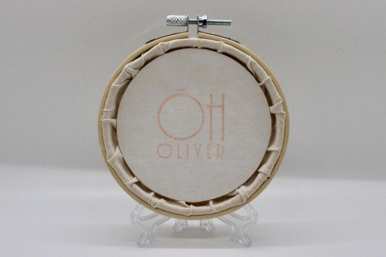 May include: Round embroidery hoop with a cream-colored fabric featuring the text "OH OLIVER" in a soft orange hue. The fabric is stretched within a wooden hoop, secured with a metal screw. The hoop is displayed on a clear plastic stand.