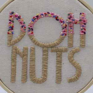 May include: Embroidered hoop art with the words "DONUTS" in a playful font. The letters "D", "O", and "H" are decorated with pink and colorful sprinkles, while the other letters are a tan color. The background is a light beige fabric.