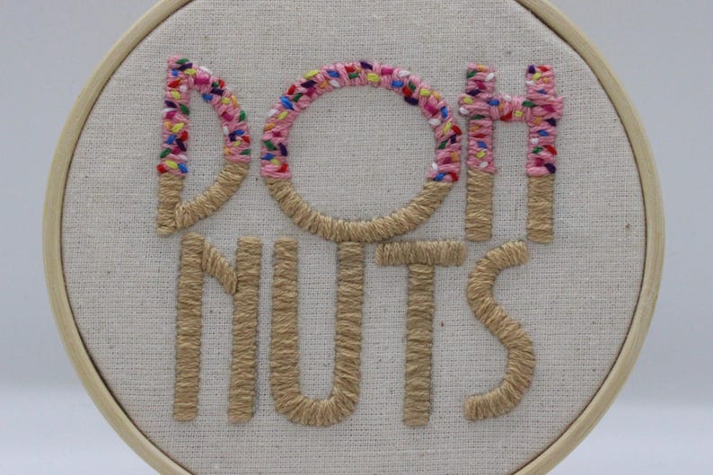 May include: Embroidered hoop art with the words "DONUTS" in a playful font. The letters "D", "O", and "H" are decorated with pink and colorful sprinkles, while the other letters are a tan color. The background is a light beige fabric.
