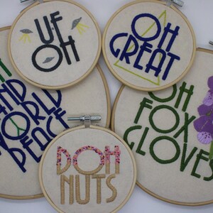 May include: A collection of embroidered hoop art with various phrases and images. The phrases include "Oh and World Peace," "Oh Great," "Oh Fox Gloves," "Doh Nuts," and "Uf Oh." The embroidery features green, blue, black, and pink thread.
