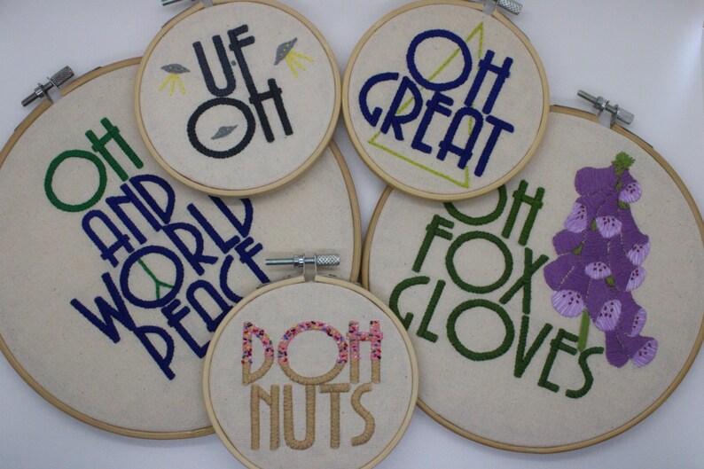 May include: A collection of embroidered hoop art with various phrases and images. The phrases include "Oh and World Peace," "Oh Great," "Oh Fox Gloves," "Doh Nuts," and "Uf Oh." The embroidery features green, blue, black, and pink thread.