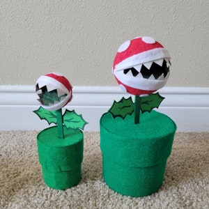 May include: Two plush, felt, red and white Piranha Plants from the Super Mario Bros. video game series. The plants are in green pots with a green felt base.