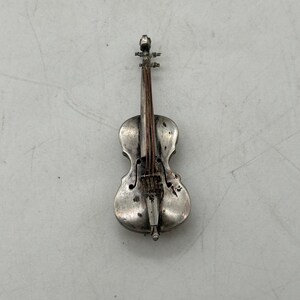 May include: A small, silver-colored violin figurine. The miniature instrument features detailed strings, a bridge, and a scroll. The violin is set against a plain, light-colored background, showcasing its intricate design.