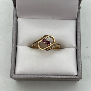 May include: A gold ring with a heart-shaped design, featuring two small, round red gemstones. The ring is displayed inside a gray jewelry box with a white interior, showcasing the ring's elegant design and color contrast.