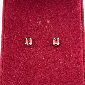 May include: A pair of small, oval-shaped, red gemstone stud earrings set in gold-colored metal. The earrings are displayed inside a red velvet jewelry box with a white satin lining.