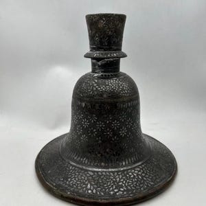 May include: An antique, dark metal bottle with a wide, flared base and a bell-shaped body. The surface is intricately decorated with floral patterns. The top has a cylindrical shape, and the overall design suggests a vintage aesthetic.