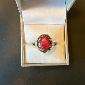 May include: A silver ring with an oval, red gemstone set in a decorative, rope-like border. The ring is displayed inside a light-colored jewelry box with a white interior, against a dark background.
