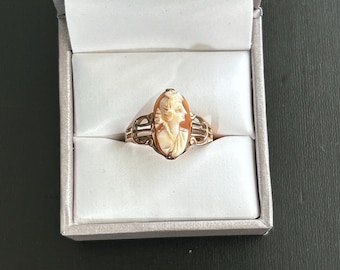 May include: Oval-shaped cameo ring featuring a carved profile of a woman in cream and orange tones. The ring has a gold-colored band with decorative filigree on the sides. The ring is displayed inside a gray jewelry box.