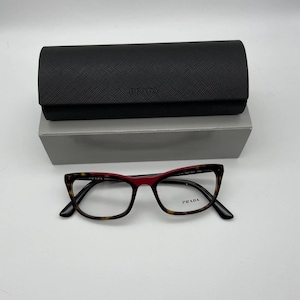 May include: A pair of Prada eyeglasses with a tortoiseshell pattern frame and red accents. The glasses are displayed with a black case and a gray box. The Prada logo is visible on the glasses and case.