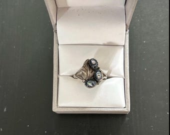 May include: A silver ring featuring a leaf-shaped metal element and three small, oval-shaped iridescent abalone shell inlays. The ring is displayed inside a light gray, hinged jewelry box with a white interior lining and a padded slot.
