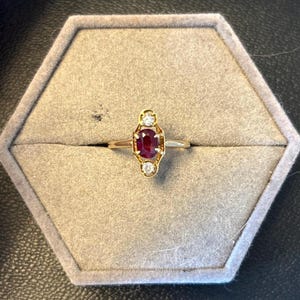 May include: A gold ring with a central oval ruby gemstone, flanked by two smaller, round, clear gemstones. The ring is set in a decorative gold setting and is displayed in a gray hexagonal ring box.
