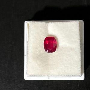 May include: A vibrant, cushion-cut red gemstone with a faceted surface, displayed centered on a square white foam insert. The gemstone exhibits a deep, saturated crimson hue and clear transparency, resting securely within the white protective display base.