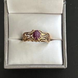 May include: A gold ring with an oval, reddish-purple gemstone at the center. The ring has an ornate, openwork design. The ring is displayed inside a gray jewelry box with a white interior.
