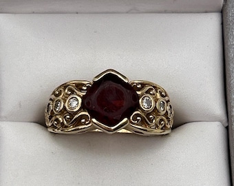 May include: A gold-toned ring featuring a large, deep red gemstone at its center. The ring's band has an ornate design with small, clear gemstones set into the sides. The ring is displayed inside a gray jewelry box.