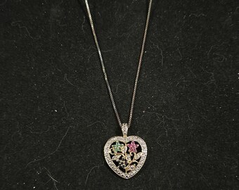 May include: A silver necklace with a heart-shaped pendant. The pendant features a floral design with green and pink gemstones, surrounded by clear crystals. The necklace is on a black background.