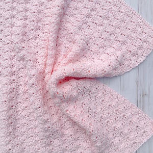 Light pink baby blanket crocheted in a shell stitch which gives it a wonderful texture.  Edges are straight with no border.  Picture taken on a white wooden background.  Blanket is laid out flat, then pulled   center on one side.