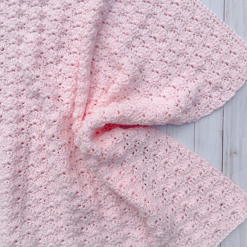 Light pink baby blanket crocheted in a shell stitch which gives it a wonderful texture.  Edges are straight with no border.  Picture taken on a white wooden background.  Blanket is laid out flat, then pulled   center on one side.